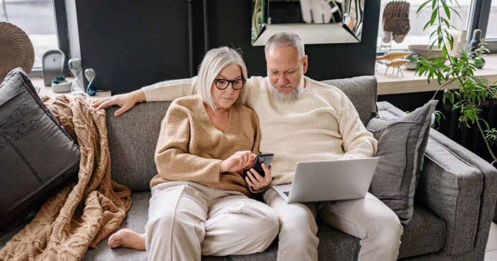 older couple looking at website with laptop and phone sitting on sofa